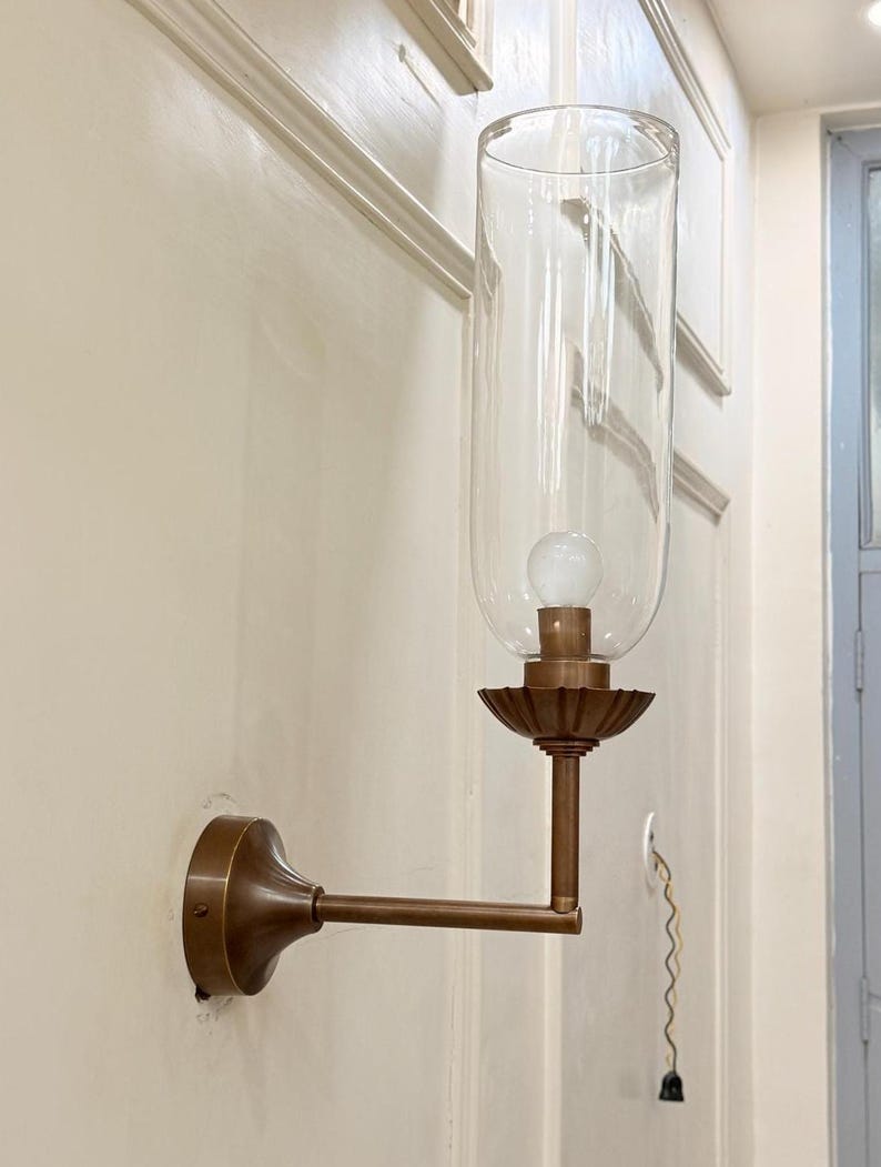 May include: A vintage-style wall sconce with a clear glass shade and a bronze-colored metal frame. The sconce features a decorative base and a long, straight arm extending from the wall. An exposed light bulb is visible inside the glass shade.