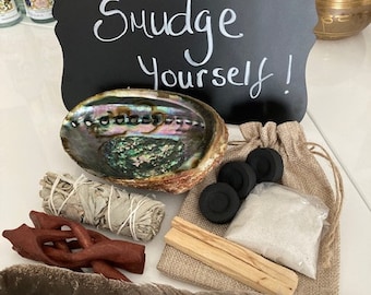 May include: Abalone shell, white sage bundle, charcoal discs, palo santo stick, and a feather. A black chalkboard with the words "Go Smudge Yourself!" is in the background. A small burlap bag contains white crystals.