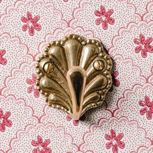 May include: A decorative brass wall hook with a shell-like design. The hook is mounted with two screws and has a curved, downward-facing hook. The background features a pink and white floral pattern.