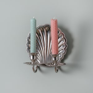 May include: A silver wall sconce in a seashell design, holding two candles. One candle is pale green, the other is pink. The candles are in silver holders. The sconce is mounted on a light blue-grey wall.