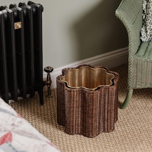 May include: A brown wicker wastebasket with a wavy top edge and a gold-colored interior. The wastebasket is cylindrical with vertical woven sides. The room has a neutral color scheme with a radiator and a wicker chair in the background.