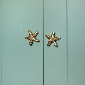 May include: Two gold-colored starfish cabinet knobs mounted on a light teal cabinet door. The knobs are detailed with small, textured bumps, and the door has vertical paneling. The knobs are a beach-themed decorative element.