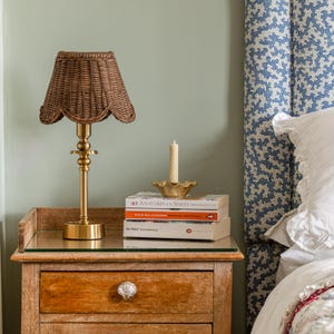 May include: A bedside table with a woven lampshade lamp, a stack of books, and a candle in a gold-colored dish. The lamp has a brass base and a scalloped shade. The books are titled "Anatomy of the Spirit" and "Robin Wall Kimmerer".