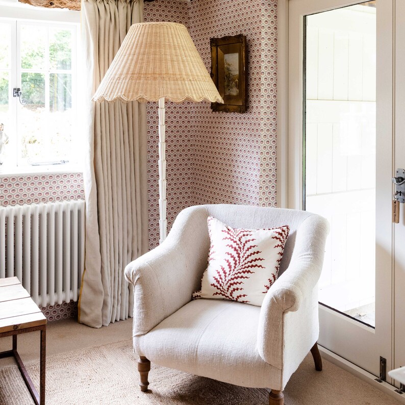May include: A white armchair with a red and white patterned pillow sits in a room with a white wicker floor lamp and a window with white curtains. The walls are covered in a pink and white patterned wallpaper.