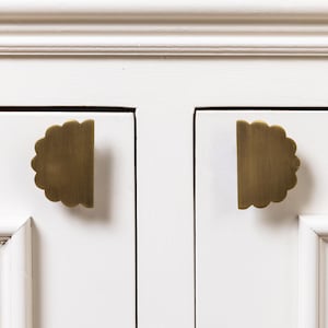 May include: Close-up of two brass-coloured cabinet pulls with a scalloped edge design. The pulls are mounted on a white cabinet door. The hardware has a half-circle shape and a matte finish.