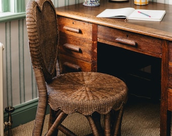 May include: A wicker chair with a heart-shaped back and a round seat. The chair is brown and has a woven design. The chair is in front of a wooden desk with drawers.