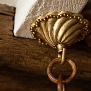 May include: Close-up of a gold-colored, ornate ceiling fixture with a shell-like design. The fixture is attached to a wooden beam and has a wooden ring hanging from it. The fixture is likely part of a chandelier or other decorative lighting.