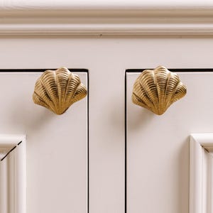 May include: Two gold-colored seashell cabinet knobs mounted on a white cabinet door. The knobs are detailed with ridges and are in a seashell shape. The cabinet door has a recessed panel design.