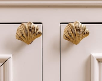 Peut inclure: Deux boutons de meuble dorés en forme de coquillage, montés sur une porte d'armoire blanche. Les boutons sont détaillés avec des nervures et ont la forme d'un coquillage. La porte de l'armoire a un design de panneau encastré.