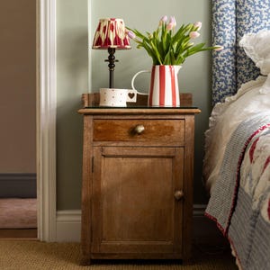 May include: A rustic wooden bedside cabinet with a drawer and door. A small lamp with a red and cream shade sits on top, beside a heart-patterned mug and a red and white striped jug holding pink tulips. The cabinet is in a bedroom.