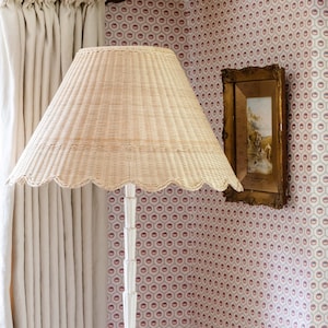 May include: A wicker lampshade with a scalloped edge sits on a white floor lamp. The lamp is in a room with white curtains and a framed picture on the wall.