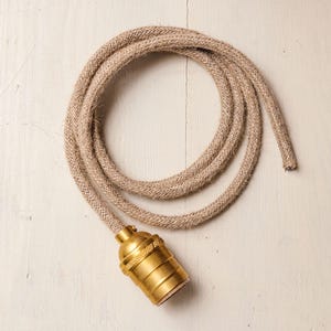 May include: A tan rope light cord with a gold-coloured bulb socket. The cord is coiled in a circular shape. The light fixture is on a white surface.