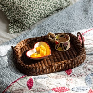 May include: A brown wicker oval tray with a heart-shaped bowl filled with yellow fruit and a colourful ceramic mug with a rope handle.