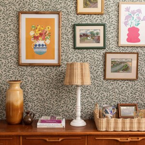 K&ouml;nnte beinhalten: Ein Wohnzimmer-Interieur mit einer gr&uuml;nen, bl&auml;ttrigen Tapete, einem Lampenschirm aus Weidengeflecht mit wei&szlig;em Fu&szlig;, einer braunen Keramikvase, einem Buch mit dem Titel "The Wild Garden" und einem Weidenkorb mit einer Keramiktasse und einem Glas.