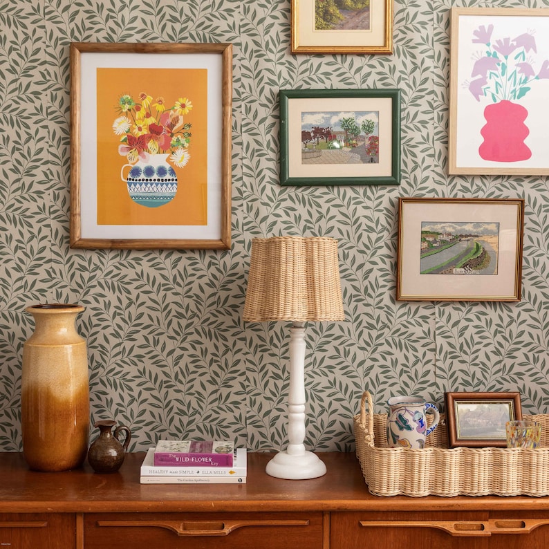 K&ouml;nnte beinhalten: Ein Wohnzimmer-Interieur mit einer gr&uuml;nen, bl&auml;ttrigen Tapete, einem Lampenschirm aus Weidengeflecht mit wei&szlig;em Fu&szlig;, einer braunen Keramikvase, einem Buch mit dem Titel "The Wild Garden" und einem Weidenkorb mit einer Keramiktasse und einem Glas.