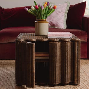 Op de afbeelding: Een bruine bijzettafel van riet met een decoratieve, golvende rand. De tafel heeft twee deuren die openen naar een opbergruimte. Een vaas met kleurrijke tulpen staat erop, samen met een boek getiteld "Patterns of India".