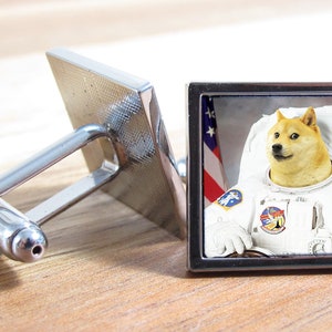 以下が含まれることがあります： アメリカの国旗を背景に、宇宙服を着た柴犬の画像が刻まれた、銀製のカフスボタンのペア。犬はカメラに向かってまっすぐ見ています。