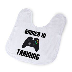 May include: White baby bib with black text that reads "Gamer in Training" and a black and green video game controller graphic.
