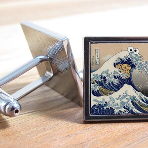 以下が含まれることがあります： Hokusai の神奈川沖浪裏をモチーフにした、正方形の画像がついた銀製のカフスボタン。クッキーモンスターがググッと目を付けて波に乗っている。