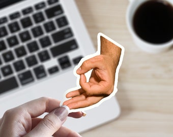 May include: A hand making the "okay" gesture, with the fingers forming a circle. The hand is on a white surface, with a laptop keyboard and a cup of coffee in the background.