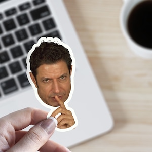 May include: A sticker of a man with dark hair and a serious expression. He is holding his finger to his lips, as if to say "shh".