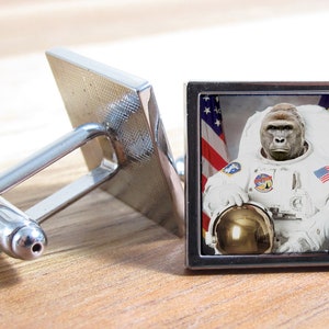 以下が含まれることがあります： アメリカの旗のパッチが腕についた白い宇宙服を着たゴリラの正方形の画像がついた銀製のカフスボタン。ゴリラは金のヘルメットを持っています。画像は赤、白、青のアメリカの旗の背景に設定されています。