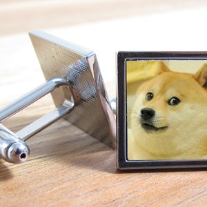 以下が含まれることがあります： 驚いた表情をした柴犬の画像が刻まれた、銀製のカフスボタンのペア。犬は右を見ています。画像は銀の枠で囲まれています。
