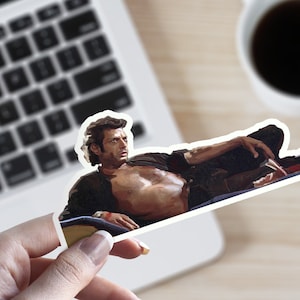 May include: A sticker featuring a man with dark hair and a black shirt, lying down with his legs crossed. He is wearing a watch on his left wrist.