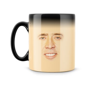 Puede incluir: Una taza de cambio de color negro y blanco con una imagen de la cara de Nicolas Cage. La taza está vacía y la imagen de Nicolas Cage es visible.
