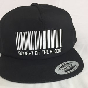 Peut inclure: Casquette de baseball noire et blanche avec un code-barres et le texte "Bought by the Blood". La casquette a une étiquette noire "The Authentic Yupoong Snapback" au dos.