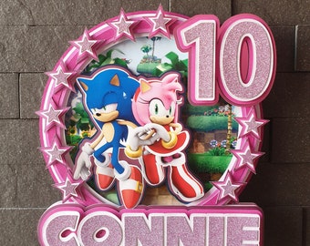 May include: Pink and blue Sonic the Hedgehog cake topper with a pink background and stars. The topper says "10" and "CONNIE".