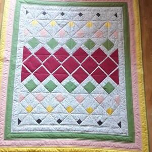 Può includere: Una trapunta patchwork con un motivo a rombi in tonalità di rosso, verde, rosa e giallo. La trapunta ha uno sfondo bianco con una stampa floreale ed è circondata da un bordo giallo, rosa e verde.