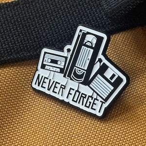 May include: A black and white enamel pin featuring a cassette tape, VHS tape, and a floppy disk. The pin reads "NEVER FORGET" in white lettering. The pin is set against a tan and black background.