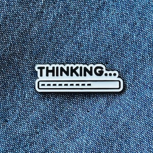 May include: A white and black enamel pin featuring the text "THINKING..." and a loading bar design. The pin is set against a textured blue background.