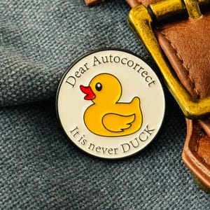 May include: A circular enamel pin featuring a yellow duck design with a red beak, set against a white background. The pin is inscribed with the words "Dear Autocorrect, It is never DUCK" around the edge.