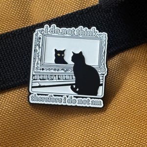 May include: A black and white enamel pin depicting two black cat silhouettes. One cat is within a picture frame, the other outside. The text "i do not think" and "therefore i do not am" are above and below the frame.