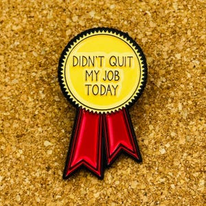 May include: A yellow and black enamel pin with the text "DIDN'T QUIT MY JOB TODAY". The pin has a circular design with a black scalloped border and two red ribbon tails.