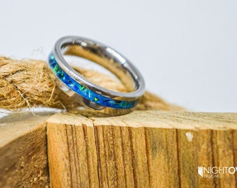 May include: A silver ring with a blue and green opal inlay. The ring is sitting on a piece of wood.
