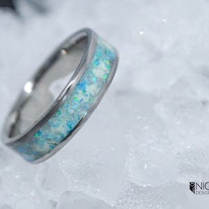Könnte beinhalten: Ein silberner Ring mit einer blauen und weißen Opal-Einlage. Der Ring liegt auf einem Hintergrund aus weißem Eis.