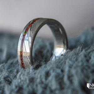 May include: A silver ring with a red and green inlay, set against a fuzzy blue background.