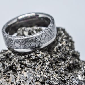 May include: A silver ring with a unique pattern, made from meteorite material. The ring is set on a dark gray rock.