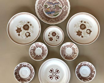 May include: A collection of vintage dinnerware featuring a variety of plates and saucers. The plates have floral and landscape designs in brown and white. The saucers have a brown patterned design. The arrangement includes large and small plates.