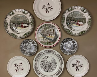 May include: A collection of decorative plates with various designs. Some plates feature winter scenes with red buildings, while others have floral or geometric patterns. The plates are in shades of white, red, green, and brown.