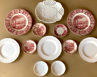 May include: A collection of 11 white and red ceramic plates with floral and landscape designs. The plates are arranged in a circle on a brown surface.
