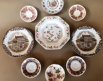 May include: A collection of eight vintage brown and white ceramic plates with floral and house designs. The plates are arranged in a circle on a beige surface.