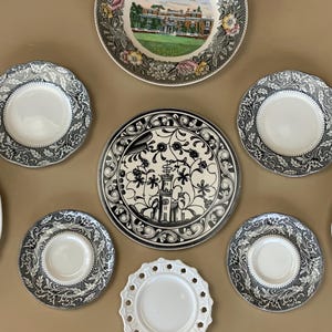May include: A collection of decorative plates in various sizes and designs. The plates feature floral, architectural, and ornamental patterns in black, white, and gray. One plate has a central image of a building.