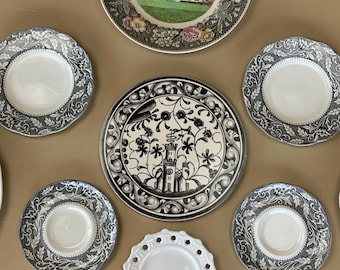 May include: A collection of decorative plates in various sizes and designs. The plates feature floral, architectural, and ornamental patterns in black, white, and gray. One plate has a central image of a building.