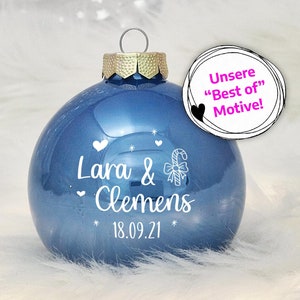 May include: A blue glass Christmas ornament with white lettering that reads "Lara & Clemens 18.09.21" and a candy cane with a bow.