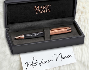 May include: A black pen with a rose gold trim and the name "Annabell" engraved on it. The pen is in a black box with the words "MARK TWAIN" on the lid. A white card with the words "Mit deinem Namen" is in front of the box.