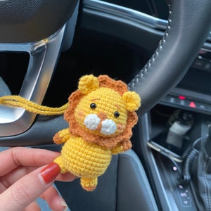May include: A crocheted lion keychain with a yellow body, brown mane, and white facial features. The keychain is hanging from a car's steering wheel.